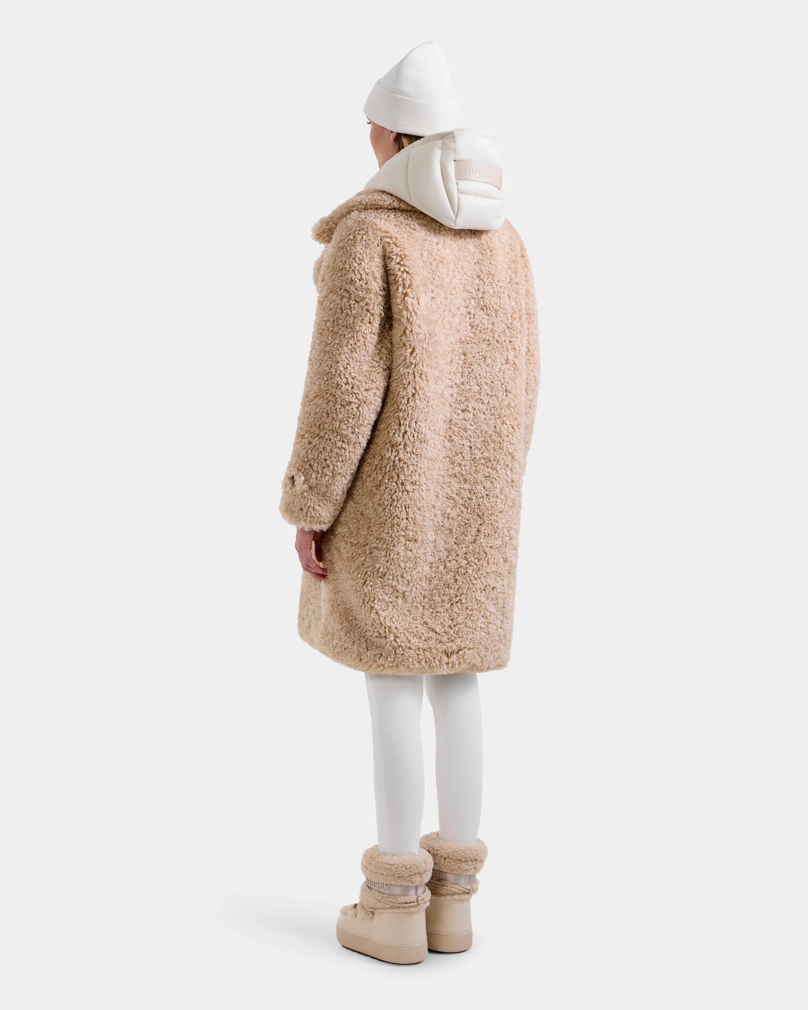 A person faces away, wearing the Rudsak EMRYN - L125588 Vanilla women’s teddy jacket with a white beanie, hoodie, leggings, and tan fur-trimmed winter boots against a plain white background.
