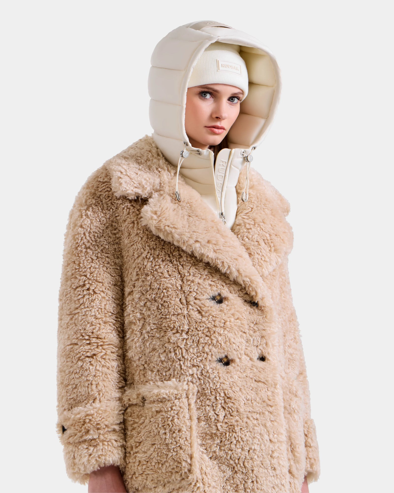 A person wears the Rudsak EMRYN - L125588 Vanilla teddy jacket, featuring large pockets and black buttons, paired with a cream puffer hood and beanie, standing against a white background.