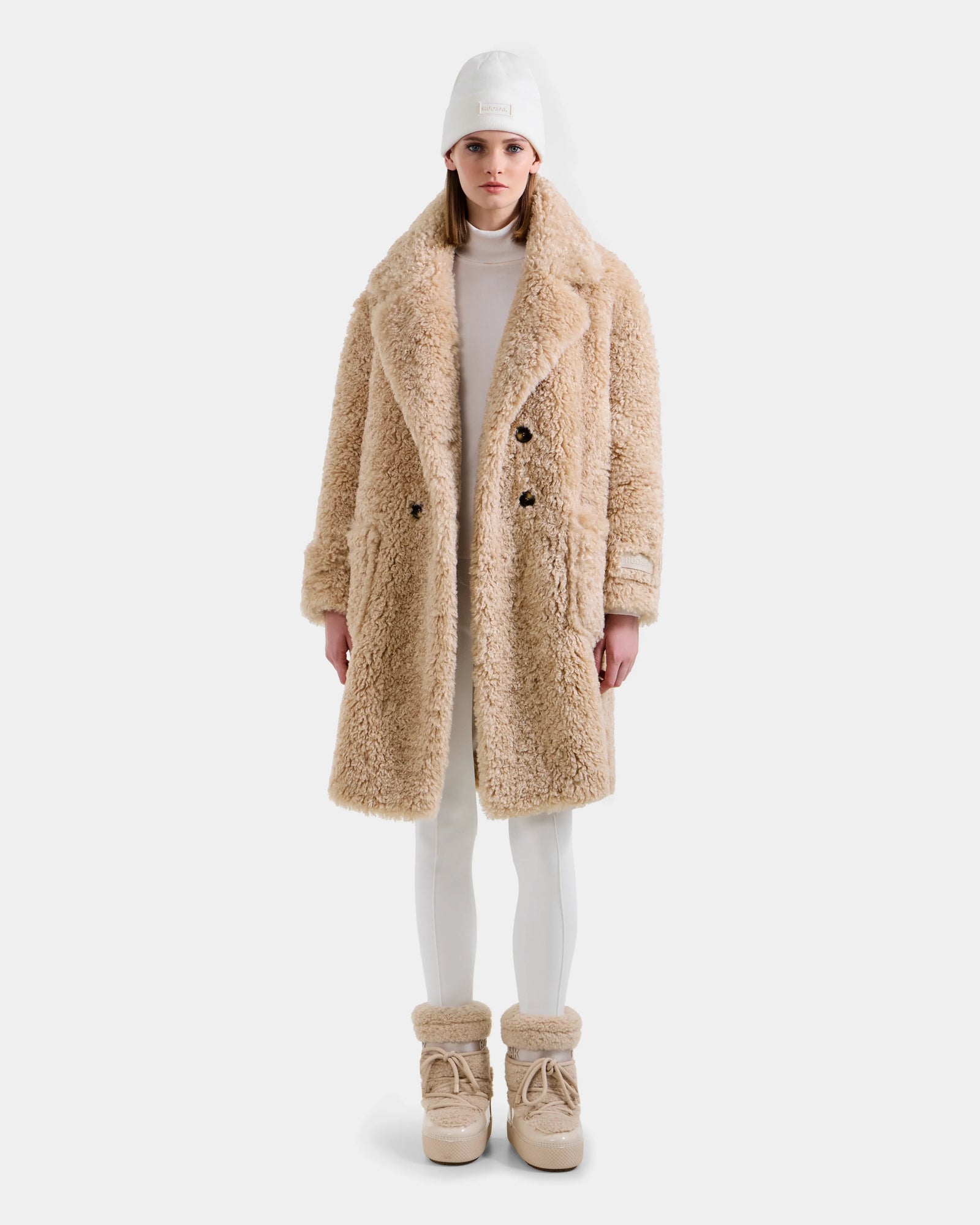 A person wearing the Rudsak EMRYN - L125588 Vanilla women's teddy jacket, white turtleneck, white pants, tan boots, and a white beanie stands facing forward against a plain white background.