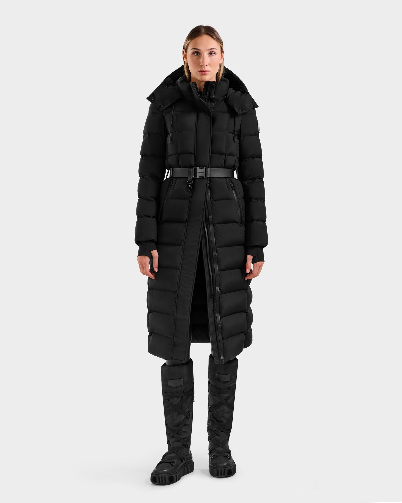 A woman faces forward against a plain light background, wearing the Rudsak FRIDA M-L - L125608 Black women’s long down puffer with a detachable hood, black pants, and matching black winter boots.