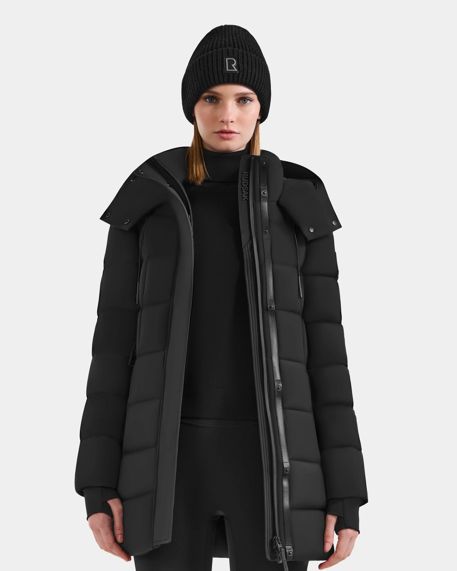 A woman models the Rudsak FRIDA M-M - L125607 Black quilted down puffer coat with a detachable hood and black beanie, standing against a white background—highlighting this winter essential.