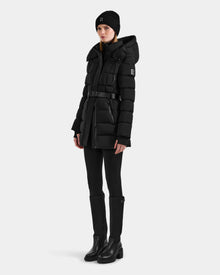 Wearing the Rudsak FRIDA M-M - L125607 Black quilted down puffer coat with detachable hood, black leggings, tall boots, and a black knit beanie, she stands against a light background—showcasing a true winter essential look.