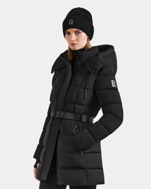 A woman wears the Rudsak FRIDA M-M - L125607 Black puffer coat with a belted waist and hood, and a matching black knit beanie, both featuring the R logo. She stands against a light gray background, facing forward.