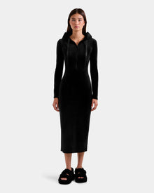 A woman faces forward in the ISABELLA - L525502 Black velour hooded dress by Rudsak, featuring a sleek midi silhouette and long sleeves, with black fuzzy slippers, set against a plain light gray background.