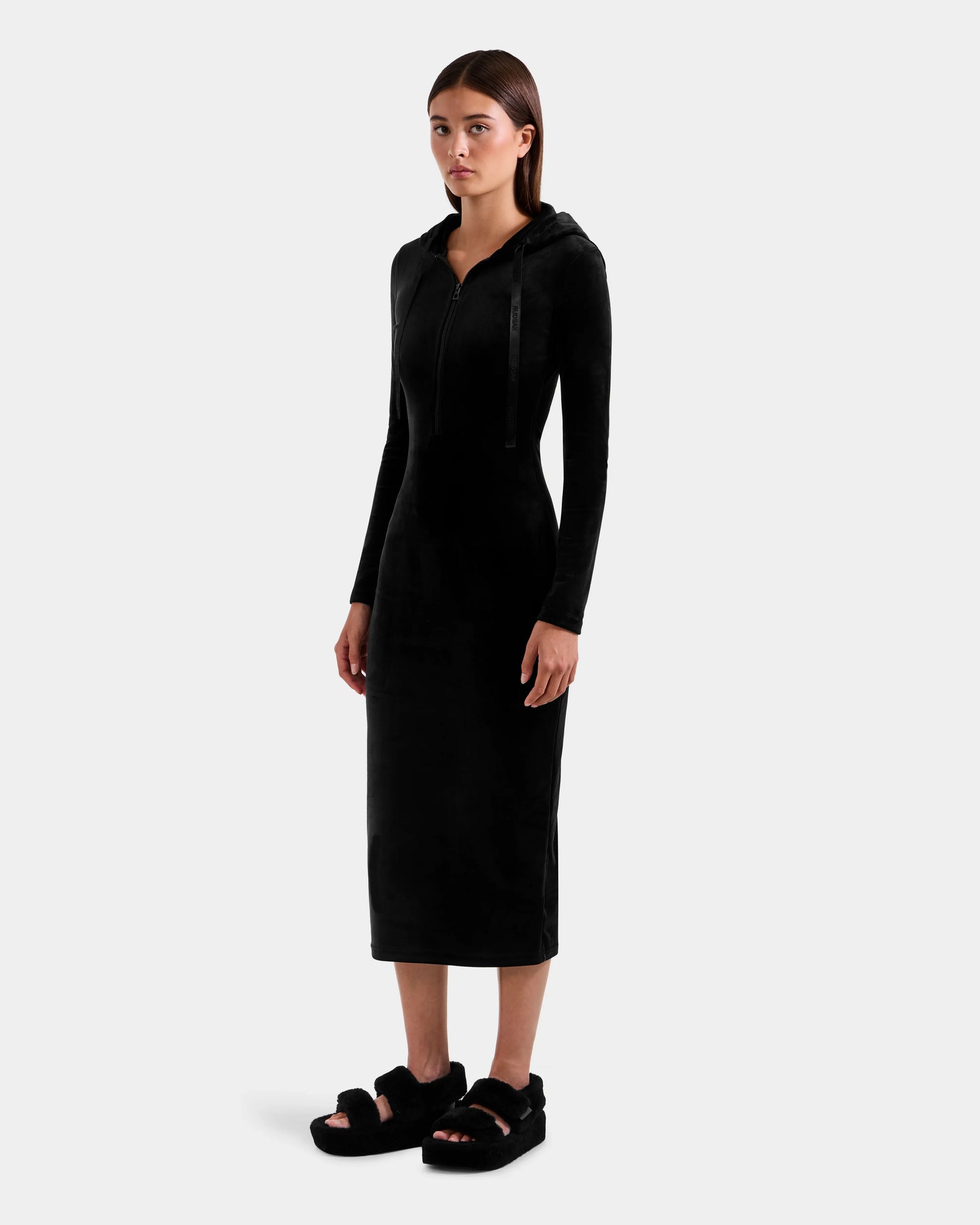 A woman with long straight hair and a neutral expression stands against a plain background wearing the Rudsak ISABELLA - L525502 black velour hooded midi dress with zipper front, paired with black fuzzy sandals.