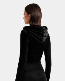 A woman with straight brown hair is seen from the back wearing the Rudsak ISABELLA - L525502 Black fitted velour hooded midi dress. The hood is up but not on her head, against a plain light gray background.