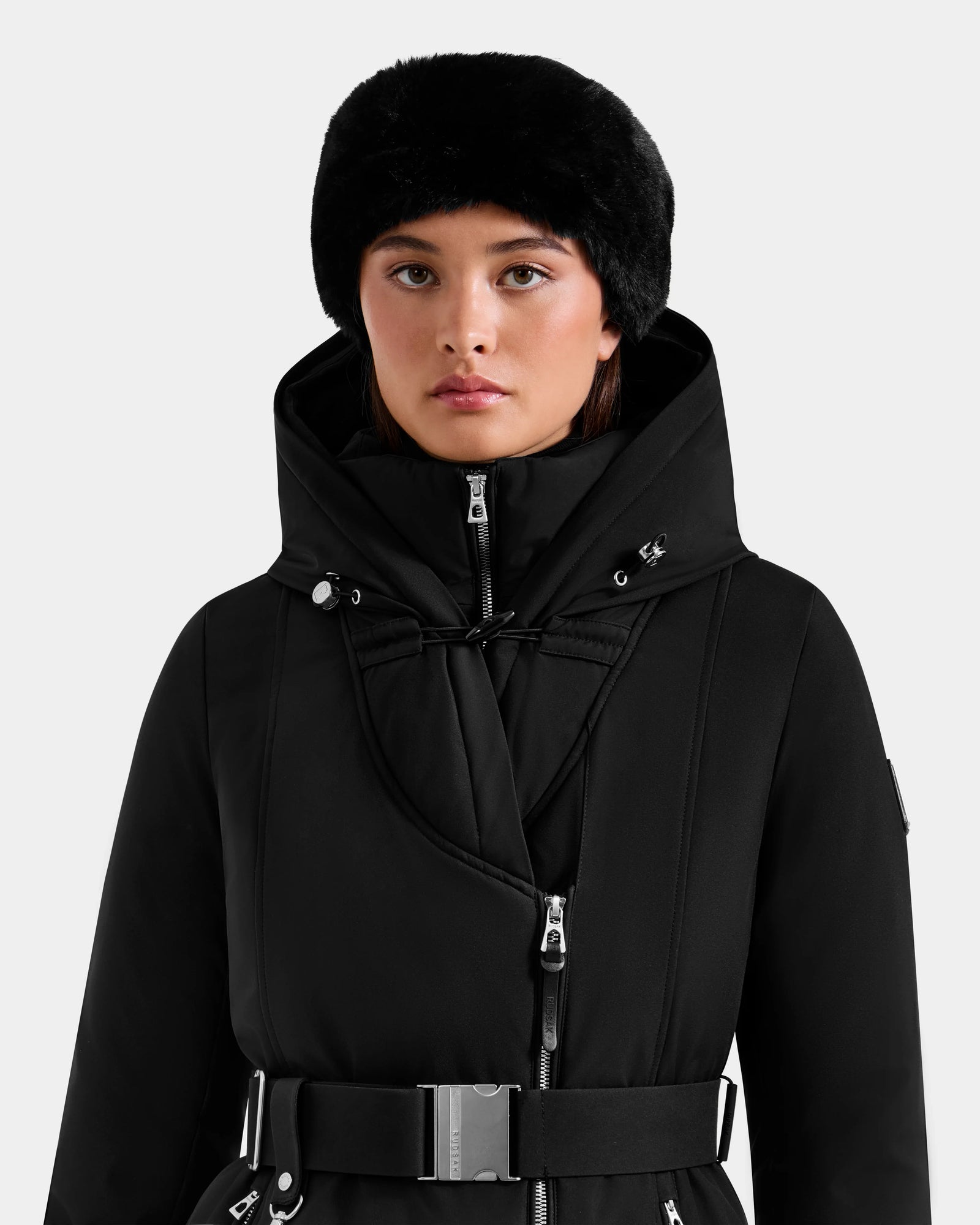 A person wearing the Rudsak ISELLE - L125547 Black women’s down parka with a belted waist and silver buckle, paired with a black faux fur hat, stands against a plain light background, looking forward neutrally.