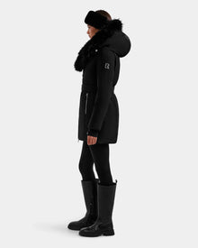 A person in profile models the Rudsak ISELLE - L125547 Black women’s down parka with a fur-trimmed hood, styled with black leggings, tall black boots, and a black headband against a plain light background.