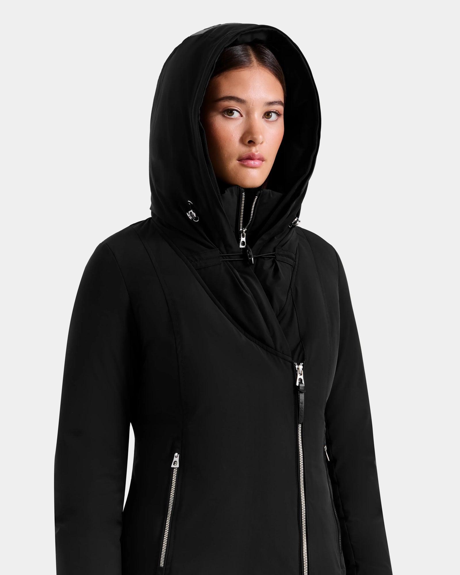 A woman models the Rudsak JANIS - L125558 Black long winter coat with silver zippers, standing slightly right and looking ahead with a neutral expression against a light gray background.