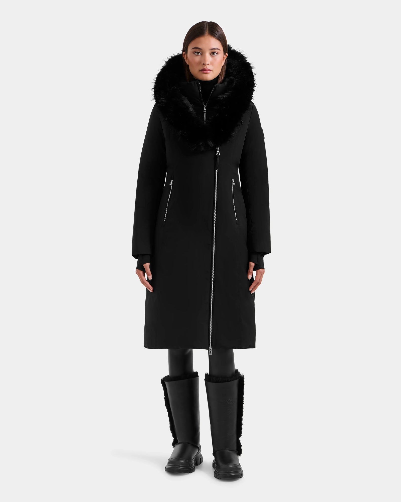 A woman faces forward against a plain light grey background, wearing the Rudsak JANIS - L125558 Black long winter coat with a large faux fur hood, paired with black leggings and fur-trimmed black boots.