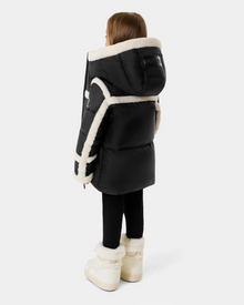 A young child stands facing away, wearing the Rudsak JULIANA K - K125550 Black puffer jacket with white sherpa trim, black leggings, and white fur-trimmed boots against a plain light gray background.