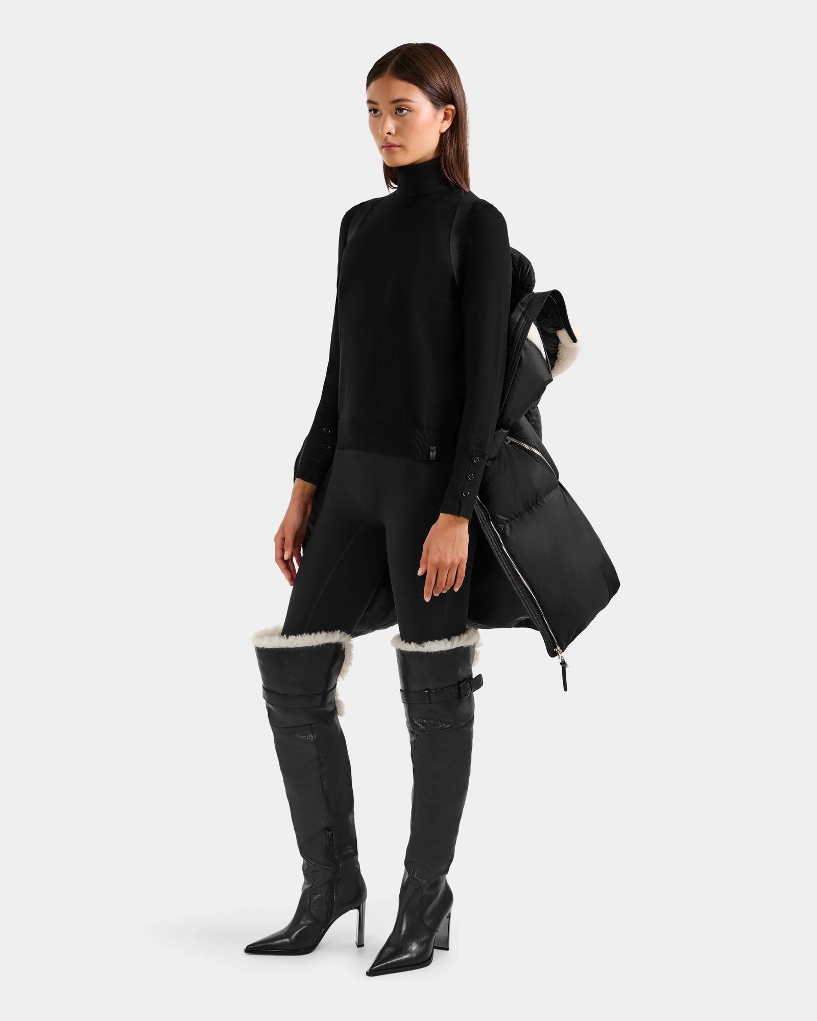 A woman stands against a white background in a black turtleneck, leggings, boots with white fur trim, and holds the Rudsak JULIANA L - L125535 Black/Vanilla women’s down puffer—ideal for chic cold-weather style.