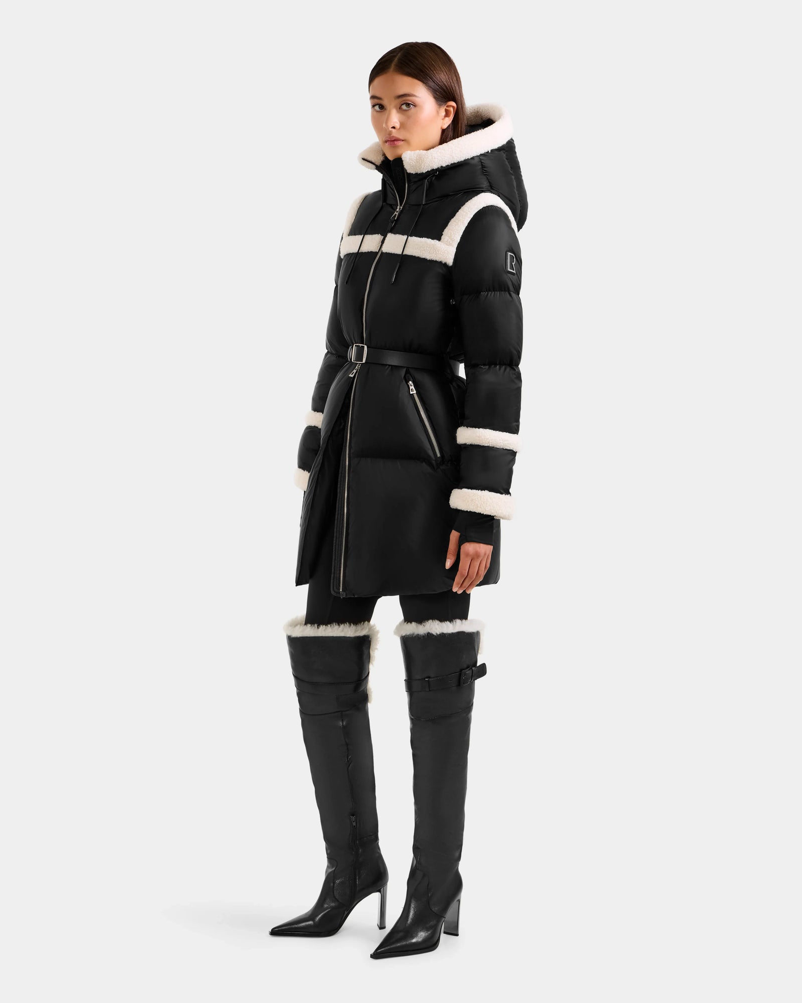 A woman wears Rudsak’s JULIANA L - L125535 Black/Vanilla: a long black belted puffer coat with white trim, hood, and zippered pockets, paired with matching black thigh-high boots also featuring white trim.
