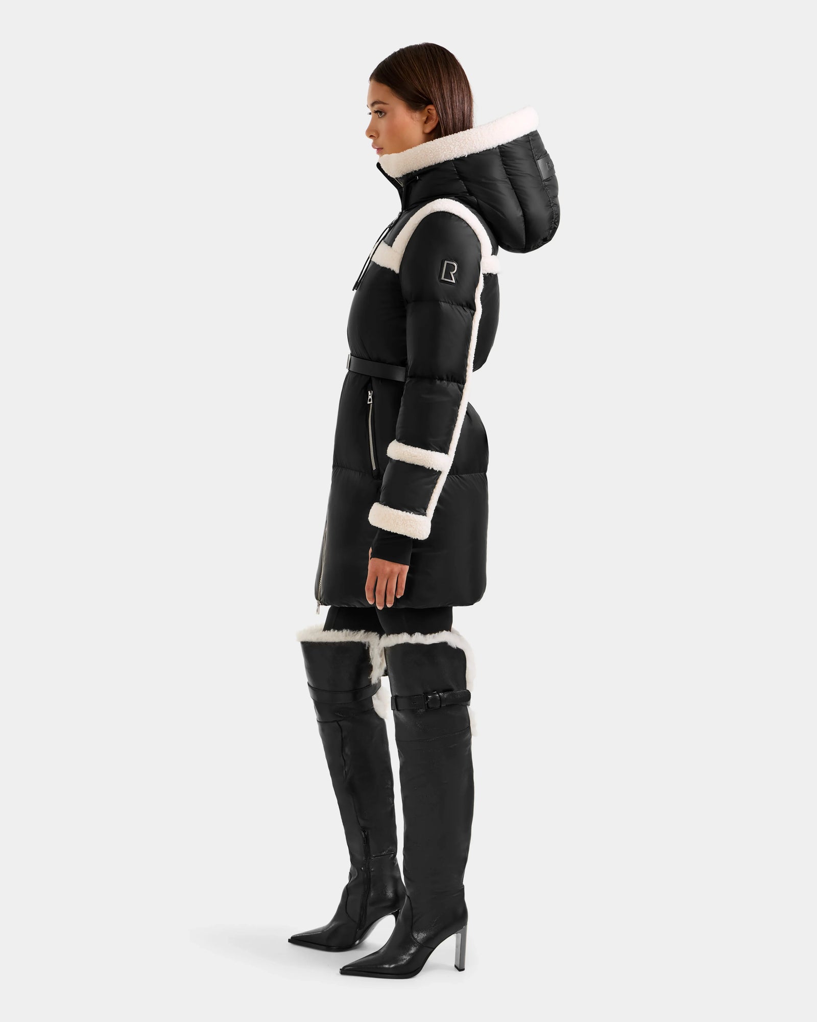A woman stands in profile wearing the Rudsak JULIANA L - L125535 Black/Vanilla, a belted women’s down puffer coat for cold weather, paired with matching over-the-knee boots with white trim against a plain light gray background.