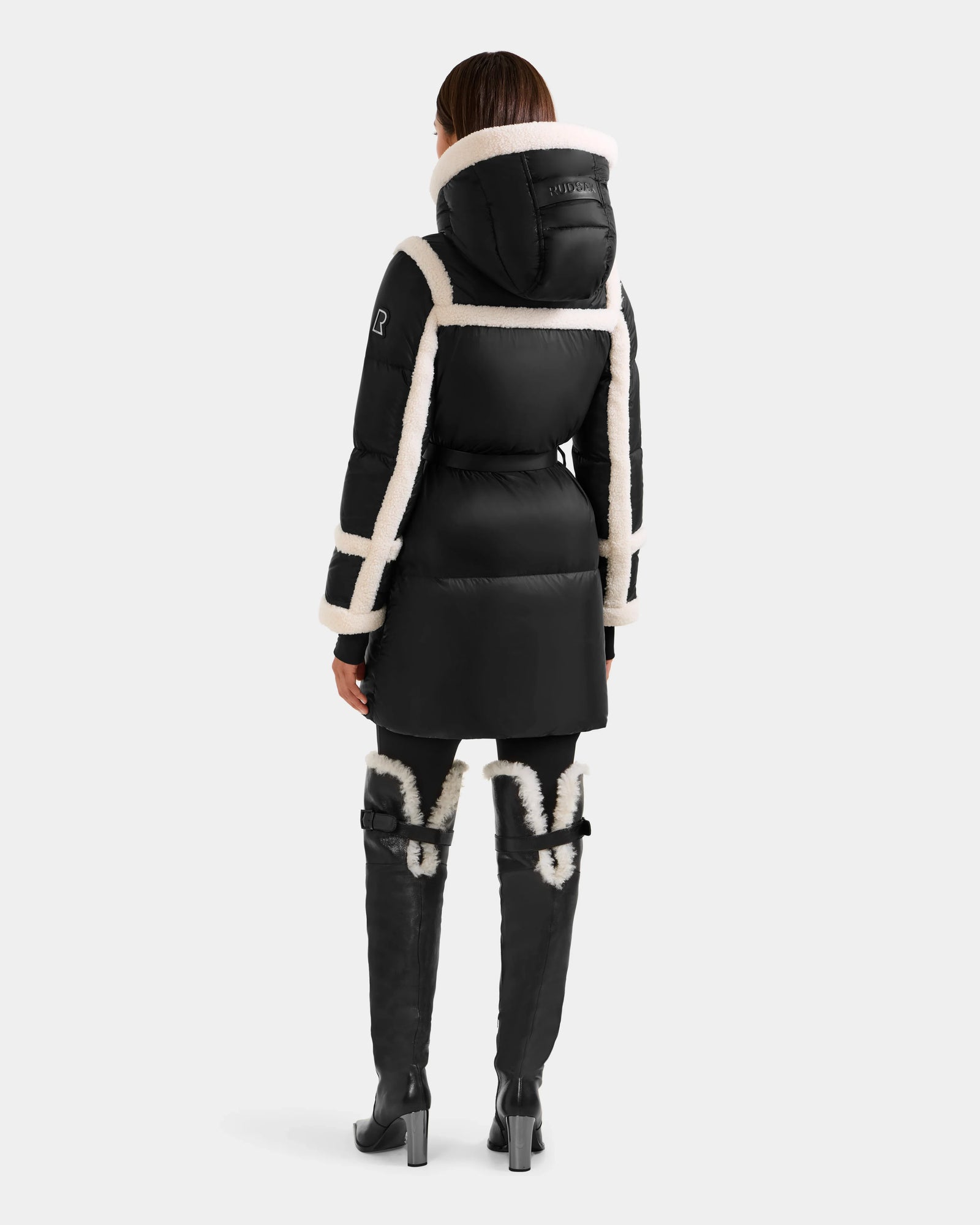A person faces away, wearing the Rudsak JULIANA L - L125535 puffer in Black/Vanilla with white faux fur trim and hood, paired with black over-the-knee boots. The cold-weather look is set against a plain light gray background.