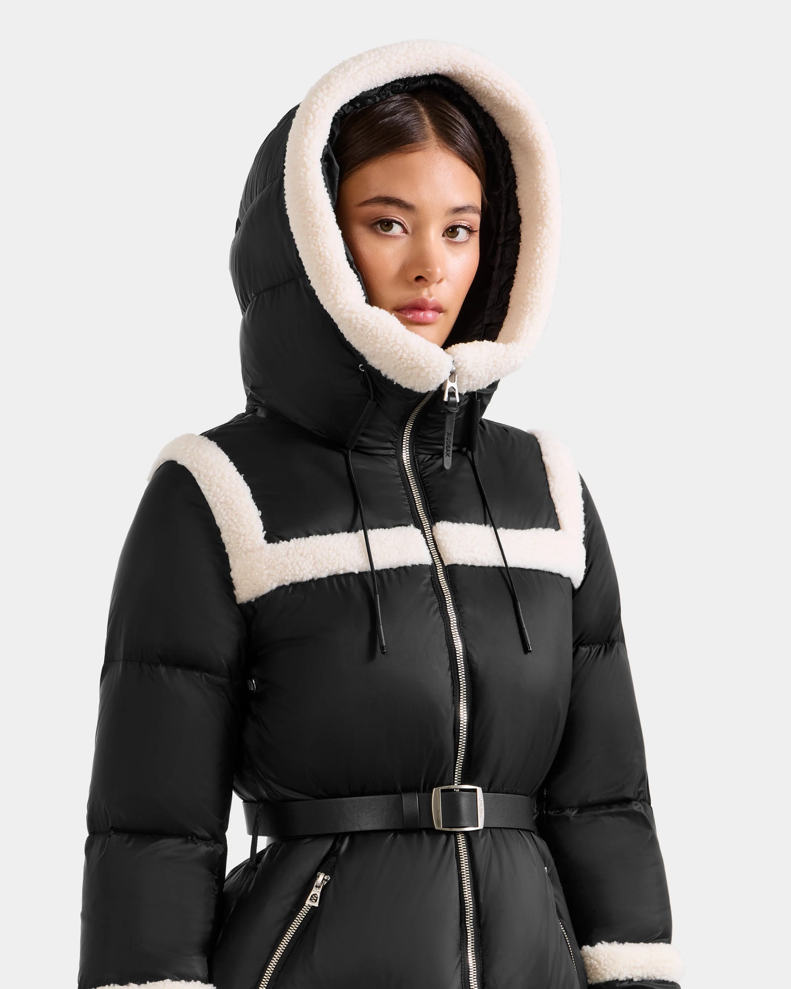 A woman wears the Rudsak JULIANA L - L125535 Black/Vanilla: a black belted puffer coat with white faux fur trim and a large hood, standing against a plain background and showcasing stylish cold-weather performance.