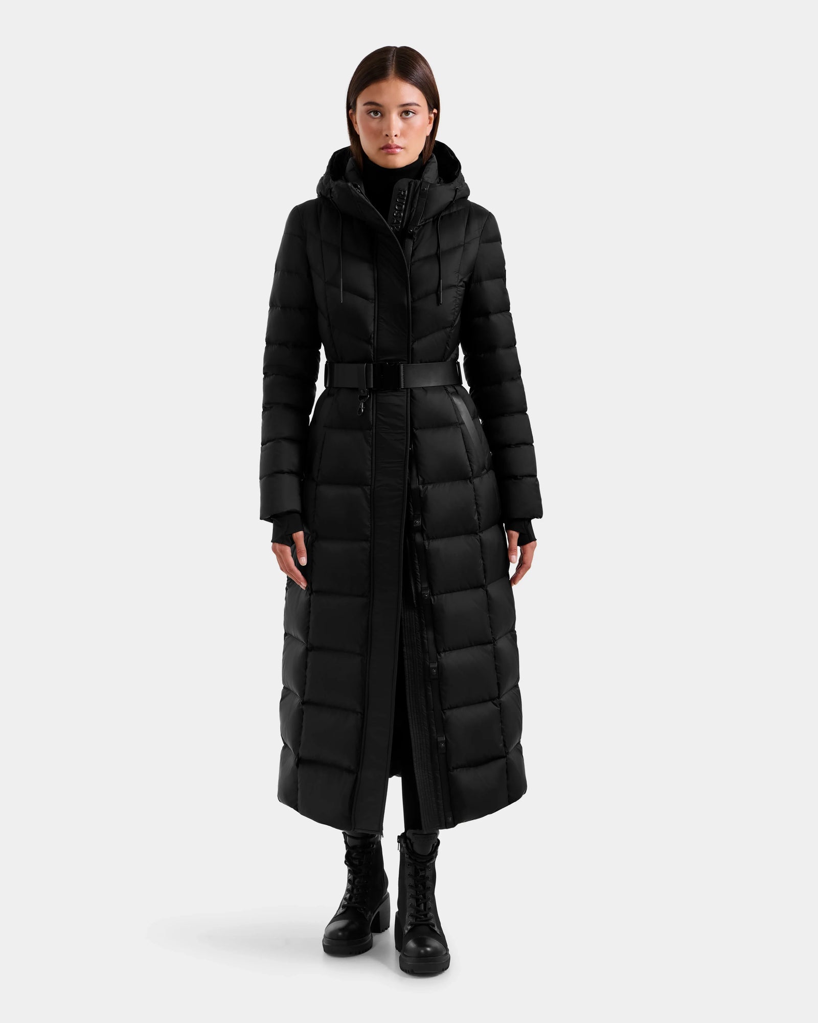 A woman faces forward wearing the Rudsak JULIETA M-L - L125559 Black maxi down puffer coat with a hood, belt, and weather-resistant shell, paired with black pants and lace-up boots. Her hands rest by her sides against a white background.