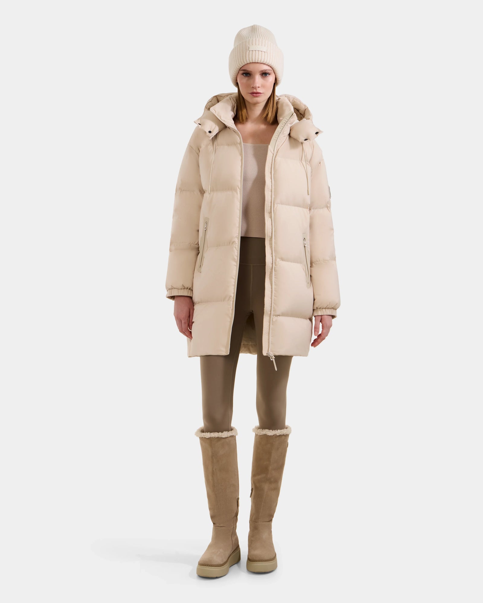 A woman wearing the Rudsak KARLEE L - 8125682 Vanilla long down puffer coat stands facing forward, styled with a beige beanie, matching top, taupe leggings, and beige fur-lined boots against a plain white background.