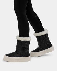 A person wears Rudsak KATHIE - L225516 Black quilted vegan leather mid boots with white faux fur trim, thick white soles, side zippers, and button details, paired with black leggings for a stylish winter look.