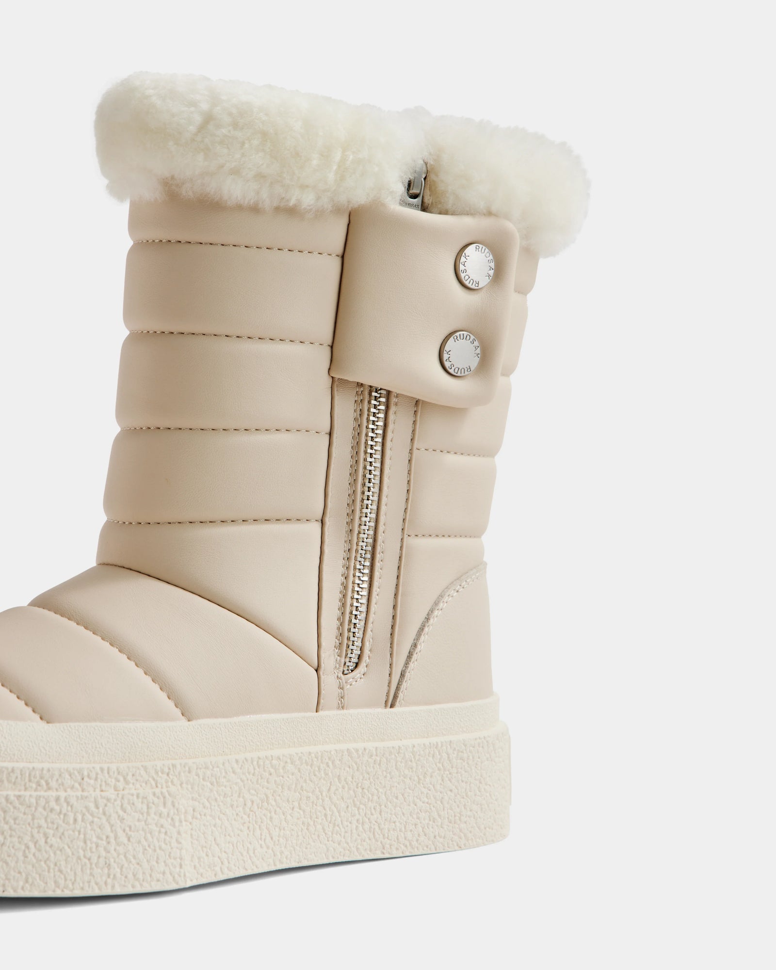 A close-up of Rudsak's KATHIE - L225516 Vanilla women's mid boot in vegan leather, featuring quilted stitching, a side zipper, metallic snap buttons, plush white faux fur cuff, and a thick textured sole.