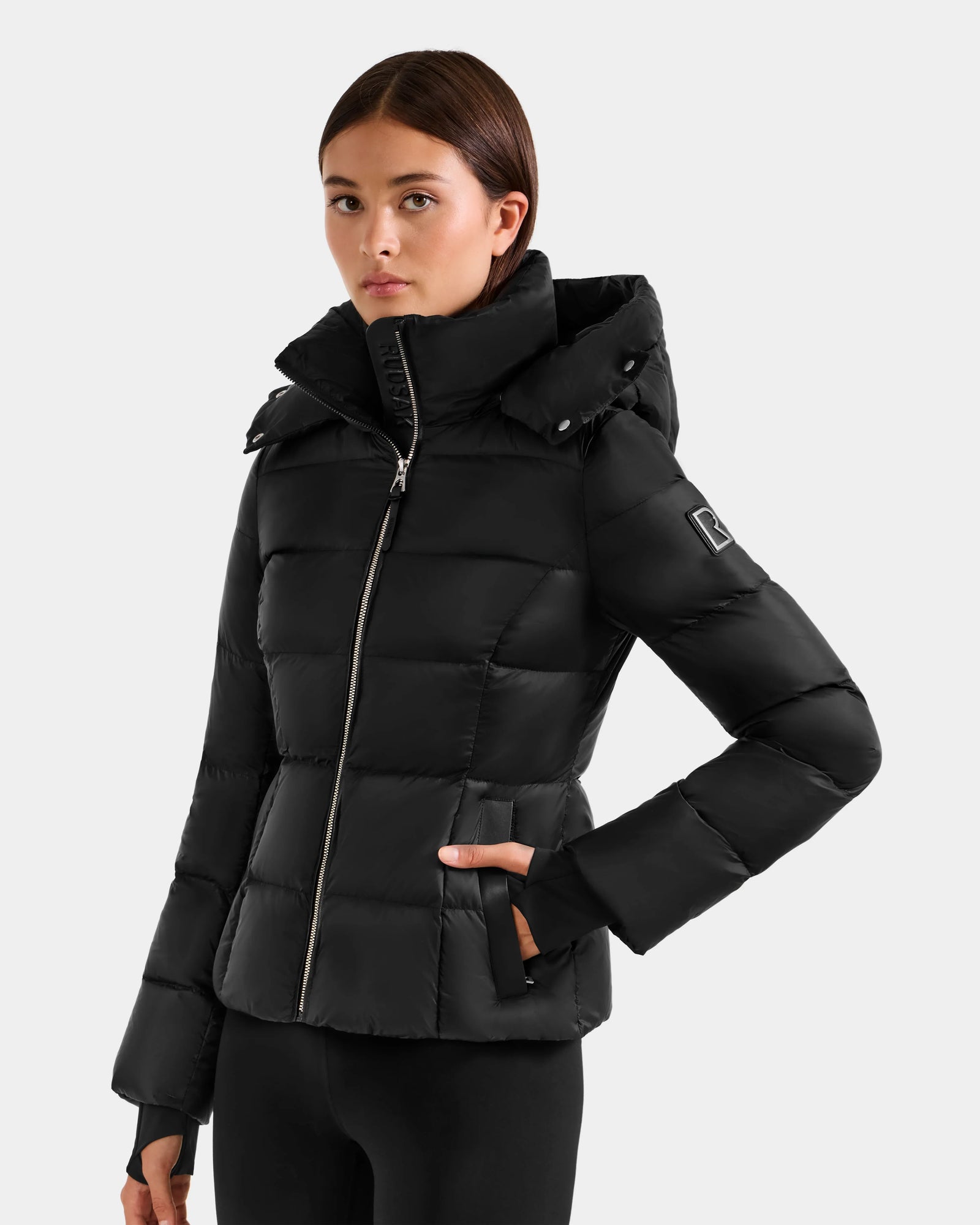 A woman models the Rudsak KATIA - L125564 Black puffer jacket with RDS-certified down, front zipper, hood, and two side pockets, standing against a plain light gray background.