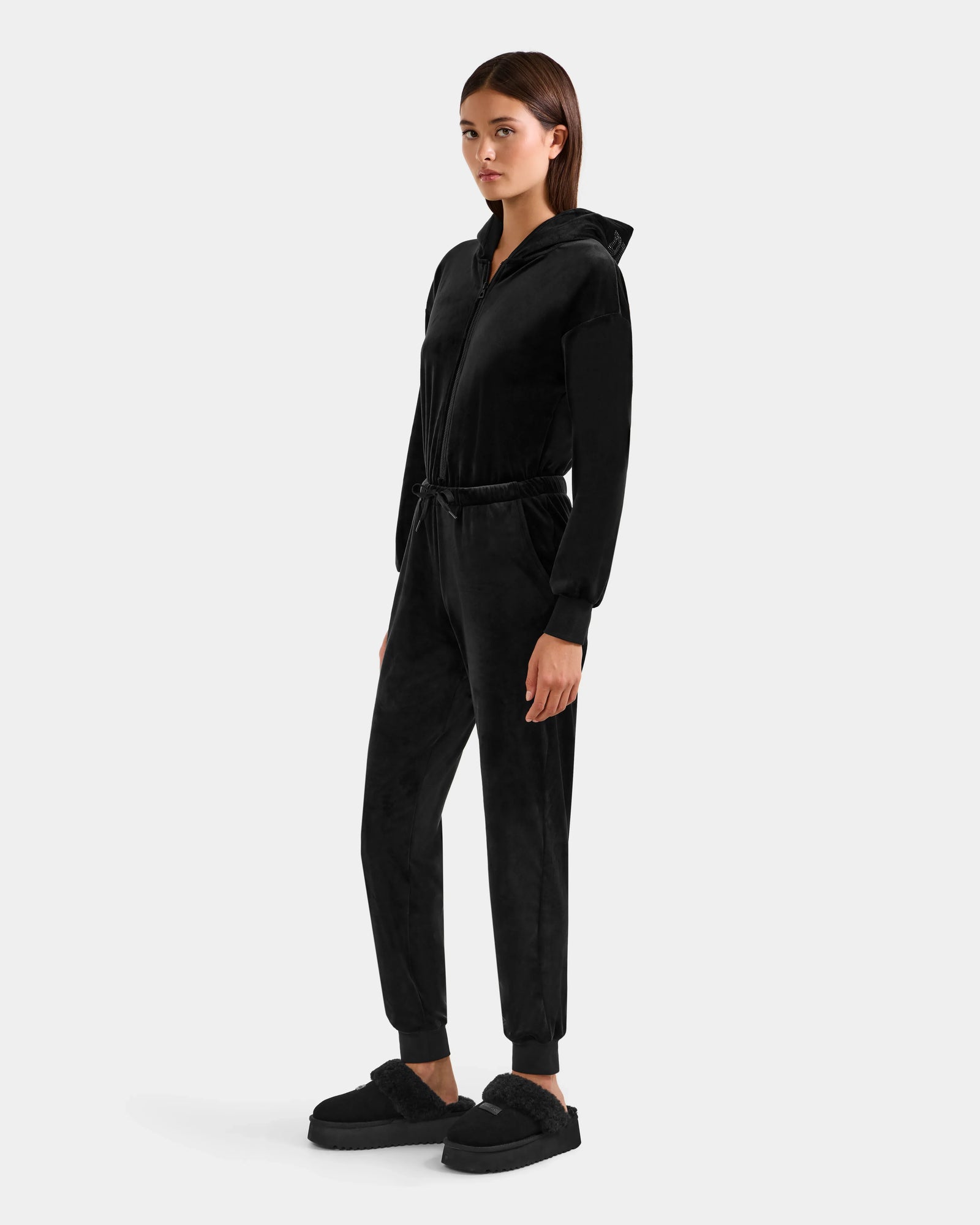 A woman stands on a white background wearing the KETZY - L525503 Black velour hooded jumper by Rudsak, featuring a drawstring waist and stretch fabric, paired with matching black slippers. She looks slightly to the side with relaxed arms.