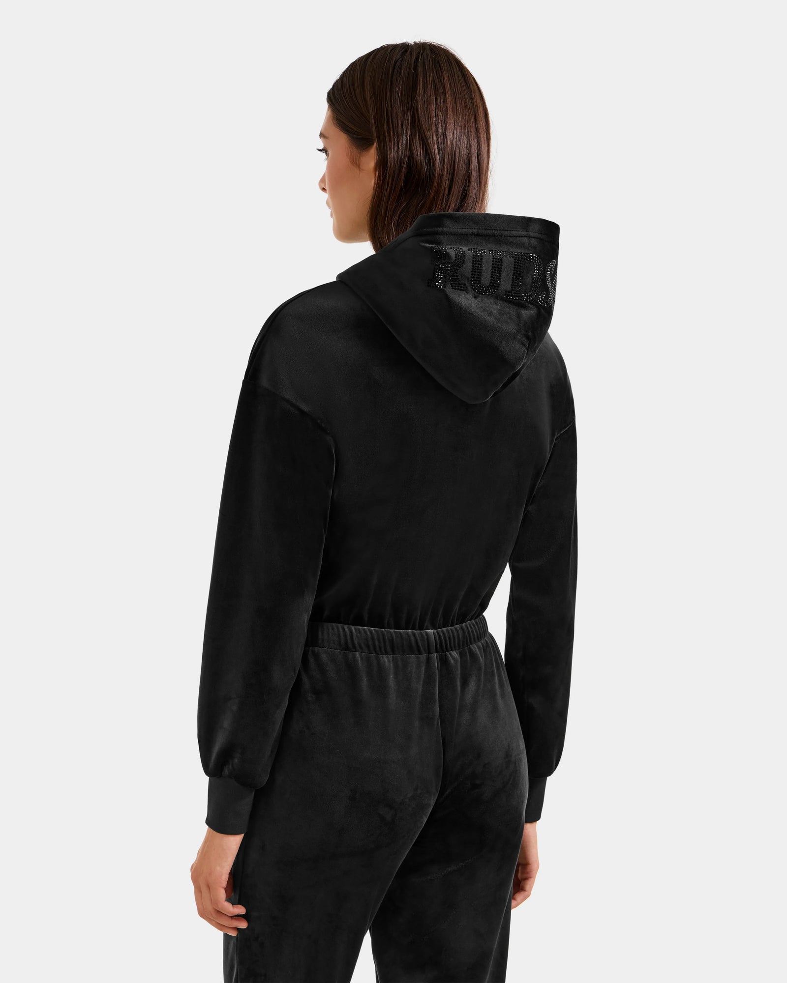 A woman stands facing away, wearing the Rudsak KETZY - L525503 Black women’s velour hooded jumper with a stretch black finish and a subtle textured design on the hood, her long brown hair falling as her arms rest at her sides.