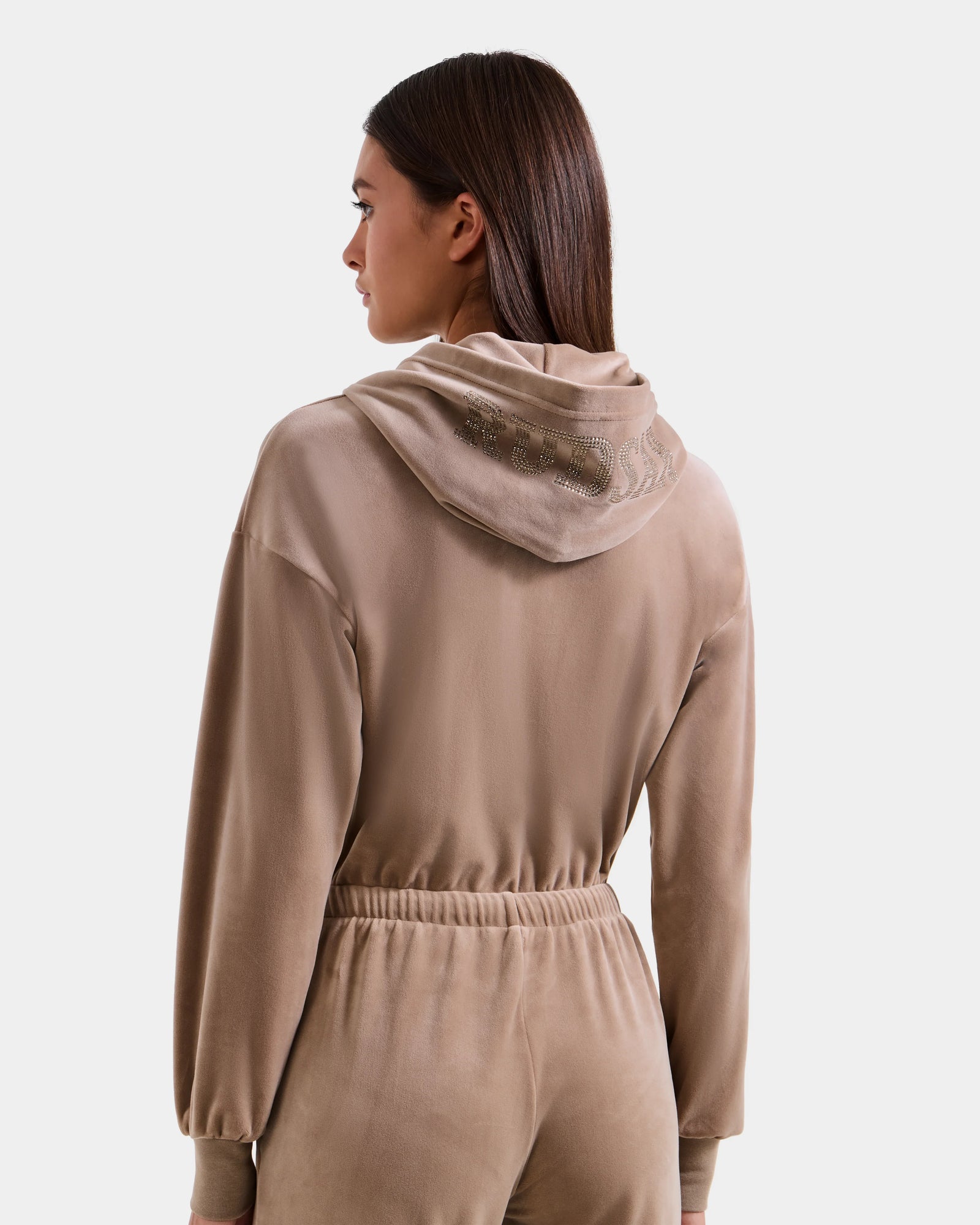 A woman with straight brown hair wears the Rudsak KETZY - L525503 Sand women’s velour hooded jumper, shown from the back, featuring crystal-sequin RUDSAK logo embellishments that add sparkle to its soft tan plush stretch velour.