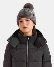 Wearing the Rudsak LANYA - L423514 Pewter puffer jacket, a woman with a high collar and a women’s pom pom hat gazes ahead with a neutral expression against a simple light background.