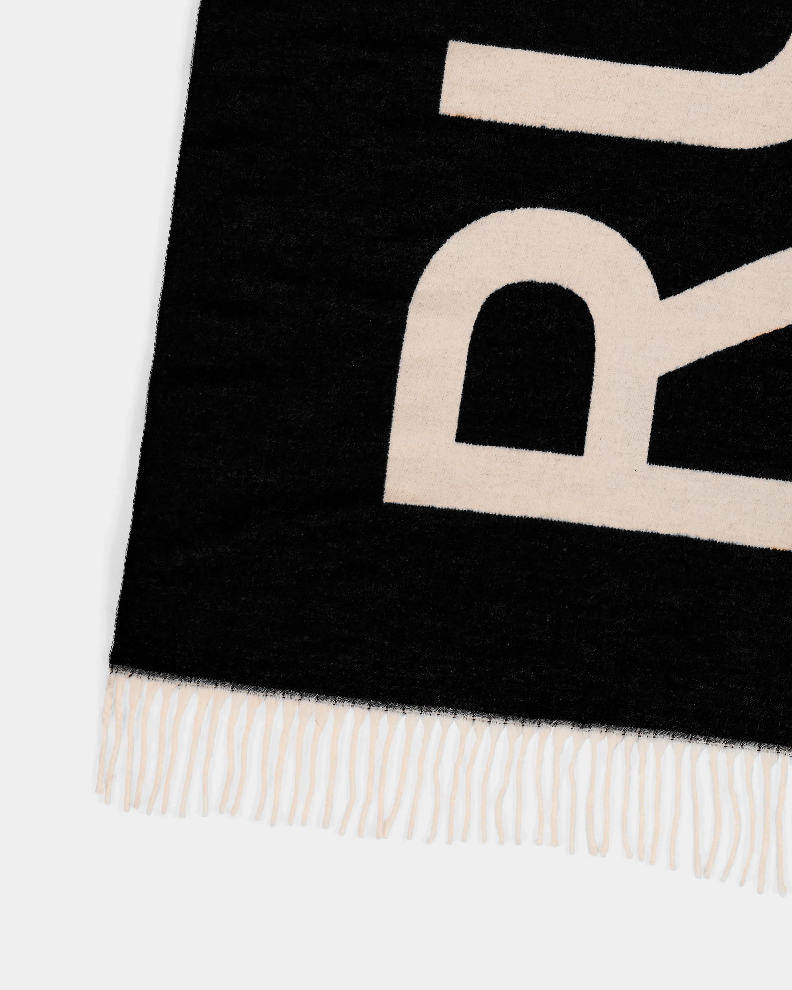A close-up of the Rudsak LEV - U425506 Black scarf, featuring bold cream letters and cream fringe. Only a portion of this woven cold-weather accessory is visible along with the lower left corner.