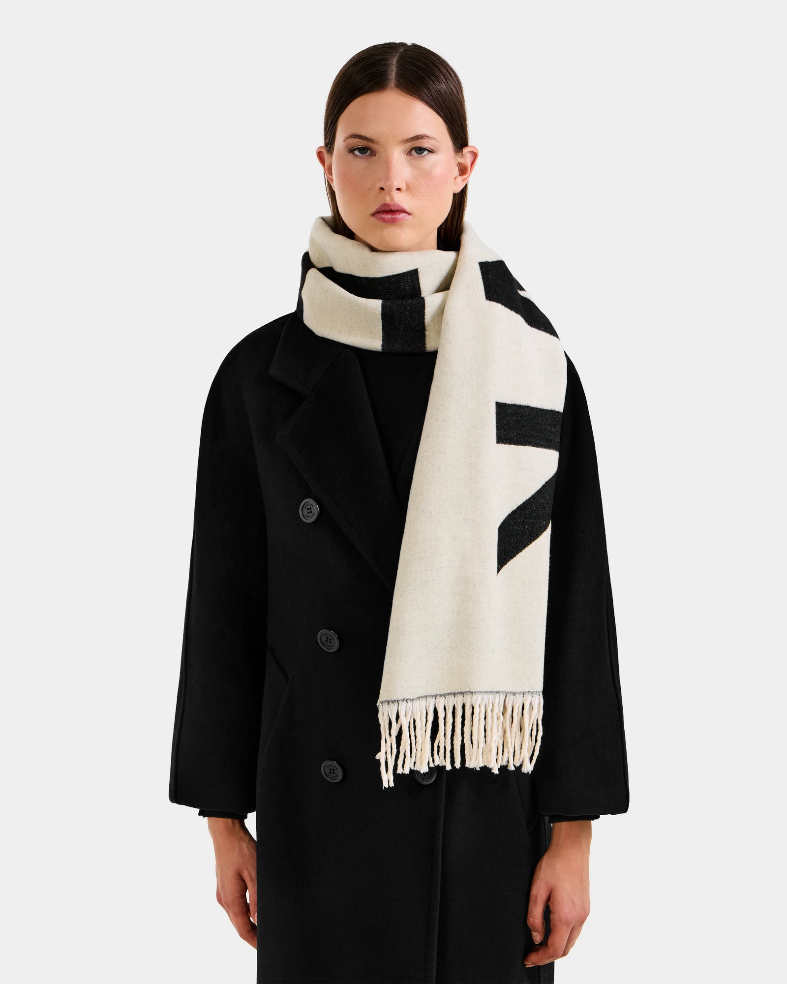 A woman wears the Rudsak LEV - U425506 Black long coat and a thick cream reversible unisex scarf with black patterns and fringed ends, posing against a plain light gray background—an elegant cold-weather look.