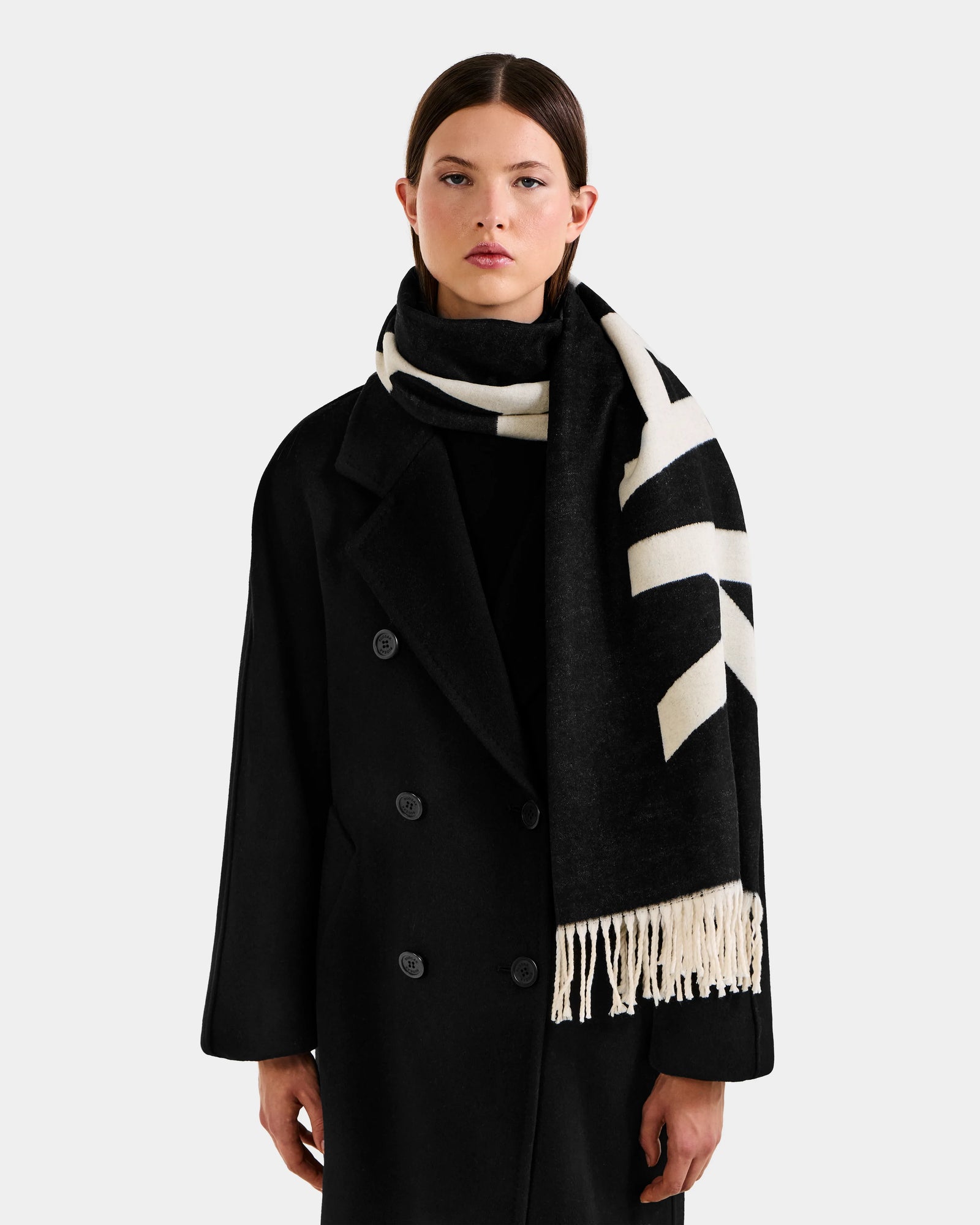 A woman wearing the Rudsak LEV - U425506 Black long coat and a reversible unisex scarf stands against a plain light background, looking at the camera. This cold-weather accessory adds style and comfort to her outfit.