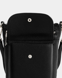 A close-up of the Rudsak LILA - L325513 Black women's crossbody bag in pebbled leather, shown with its front flap open to reveal a magnetic snap closure and a small logo visible on an item inside.
