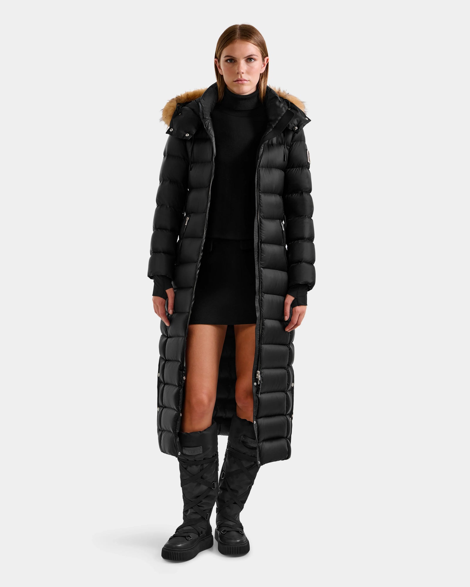 A woman wears the Rudsak LILITH L - L125528 Black puffer coat with a fur-trimmed hood, paired with a black turtleneck, black mini skirt, and knee-high black quilted boots, standing against a light gray background.
