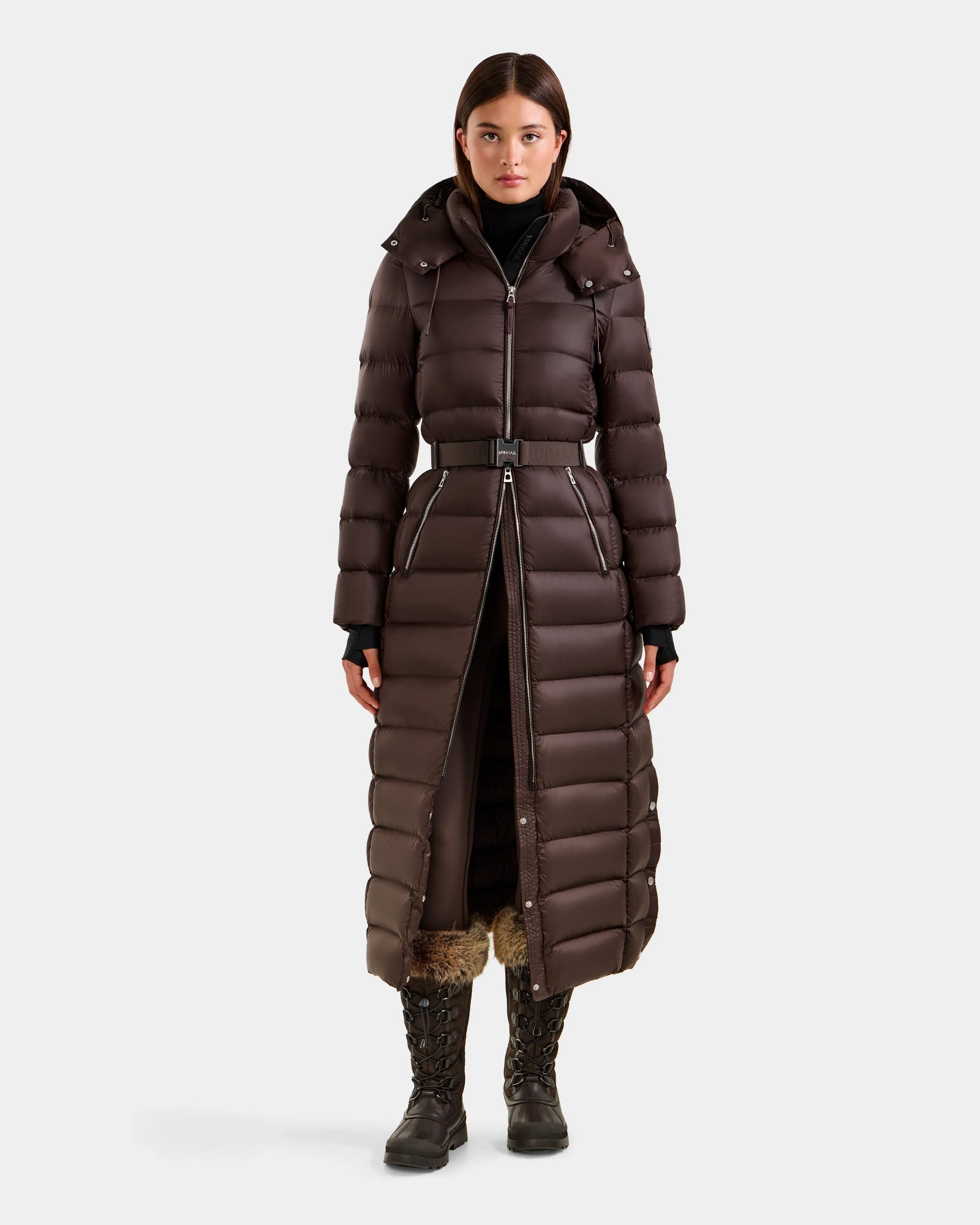 A woman faces forward in the Rudsak LILITH L - L125528 Espresso, a dark brown, RDS-certified maxi puffer with hood and removable belt. The calf-length coat features a front zipper and is styled with black lace-up boots on a white backdrop.