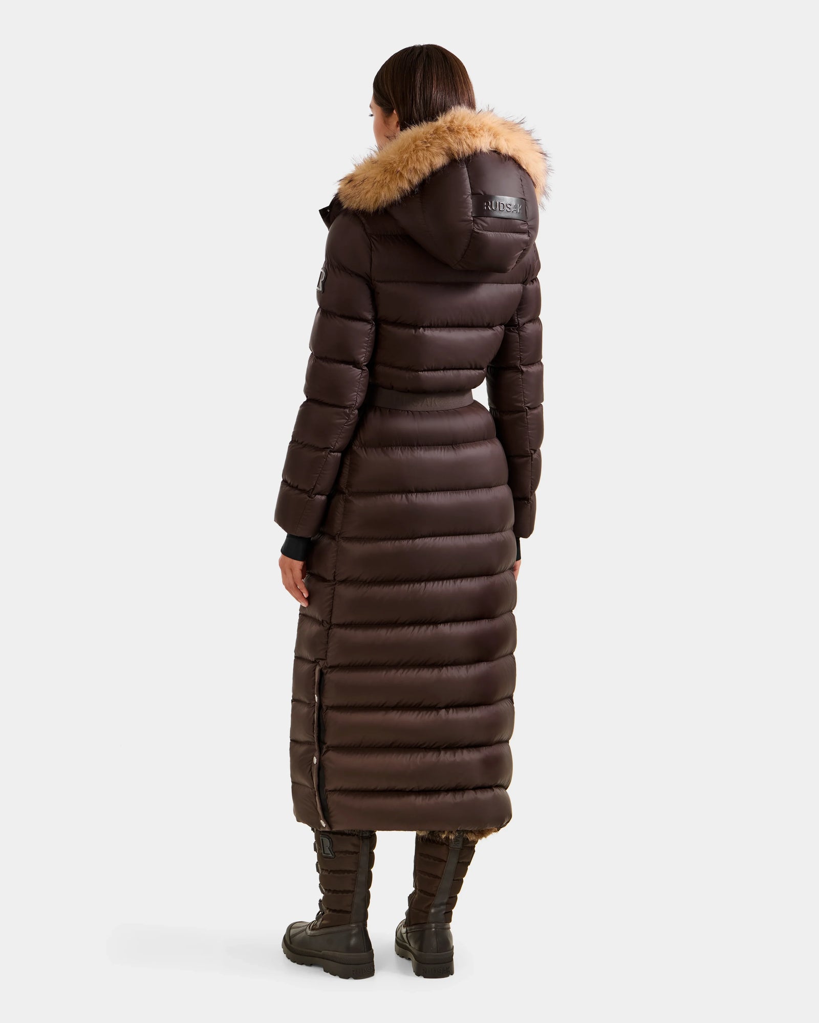 A person wearing the Rudsak LILITH L - L125528 Espresso maxi-length down puffer coat with a fur-trimmed hood stands facing away, displaying the back of the outfit against a plain light background.