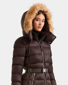 A woman wears the Rudsak LILITH L - L125528 Espresso maxi down puffer with a removable belt and light brown faux fur-trimmed hood, standing against a plain gray background.