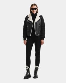 A woman in black sunglasses, the Rudsak MALA - 8125628 Black cropped shearling biker jacket, a black turtleneck, pants, and black lace-up boots with white fur lining stands against a plain white background.