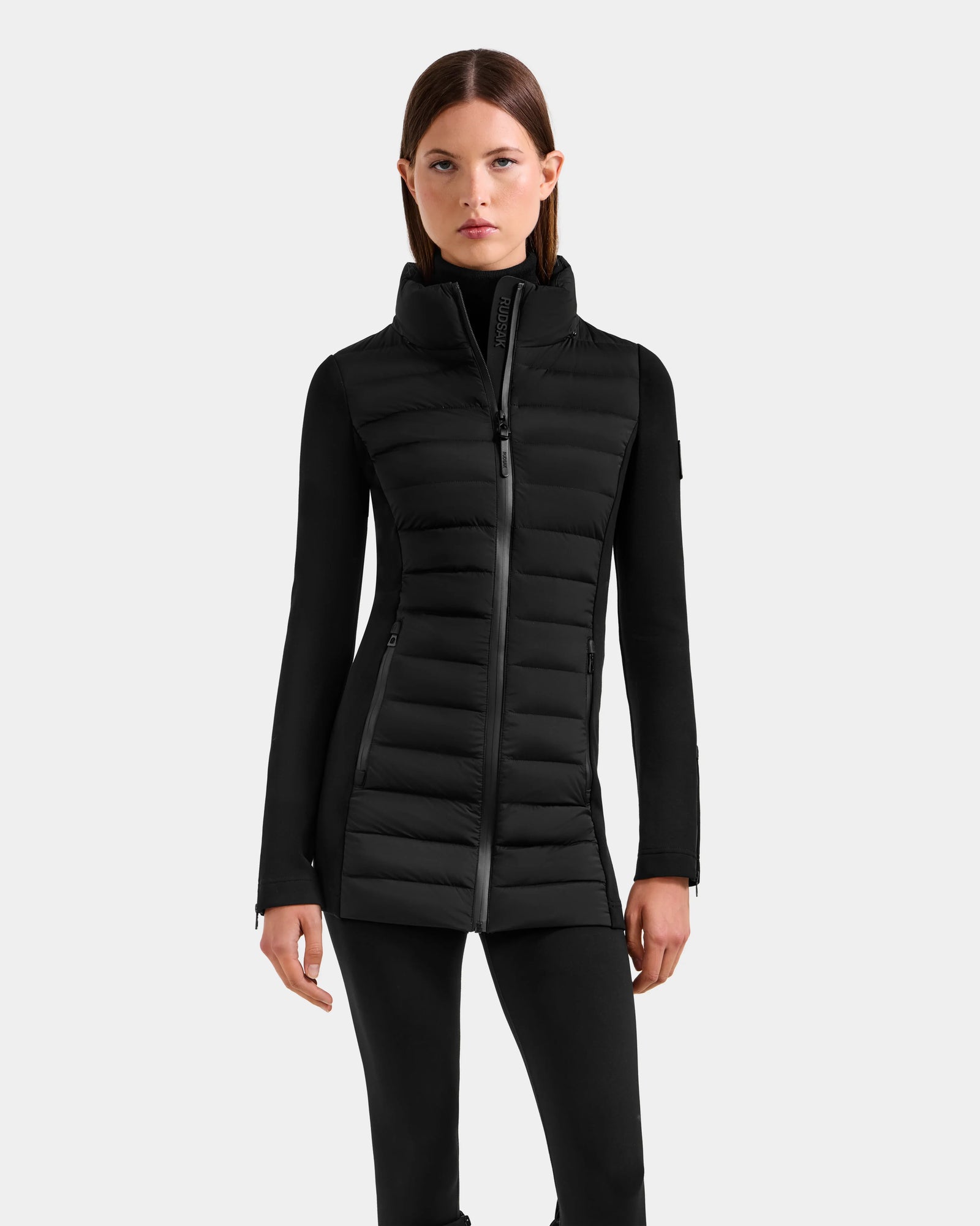 A woman stands against a light gray background wearing the Rudsak MARRY M - L125519 Black women’s mid-length hybrid coat with a high collar, layered over a fitted black long-sleeve top and black pants.