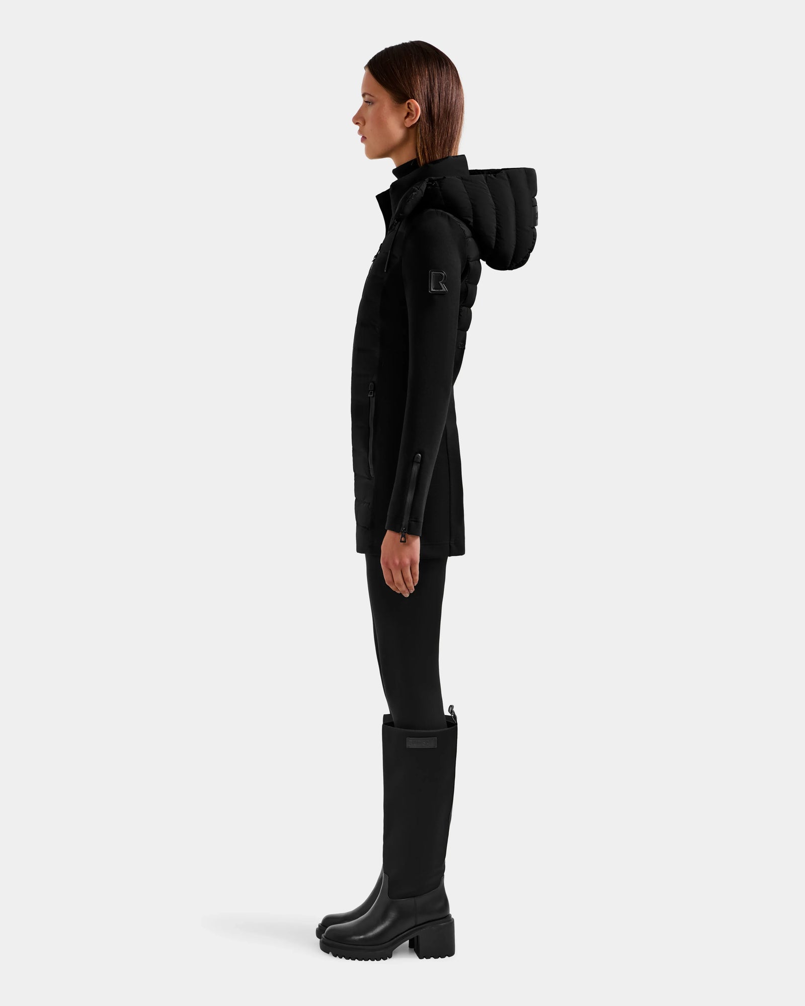 A woman stands in profile against a plain background, wearing the Rudsak MARRY M - L125519 Black women’s mid-length hybrid coat with a hood, paired with black leggings and knee-high black boots.