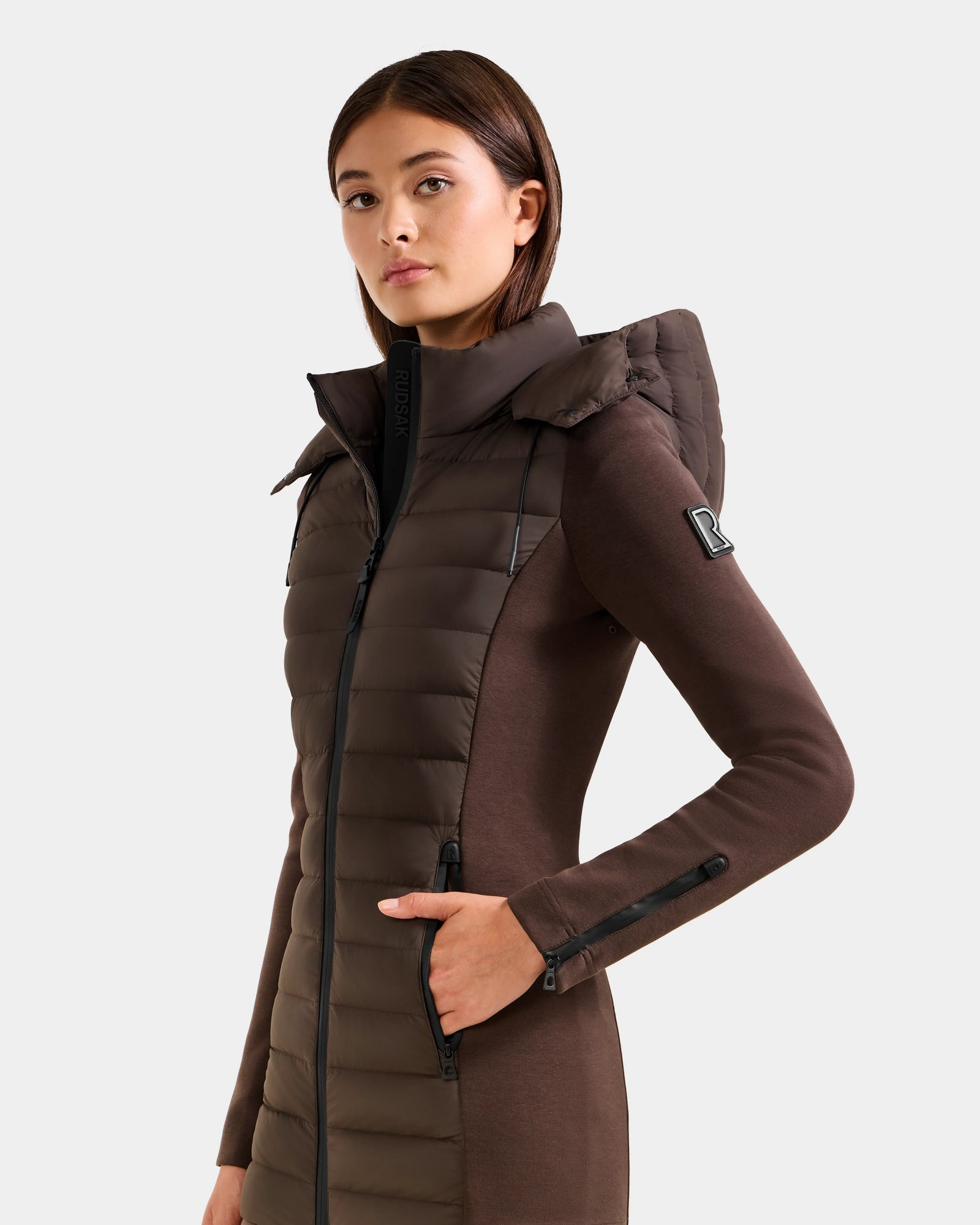 A woman wears the Rudsak MARRY M - L125519 Espresso, a brown fitted women’s mid-length hybrid jacket with a padded front, large removable hood, and zippered pockets. She stands against a plain light background facing the camera confidently.
