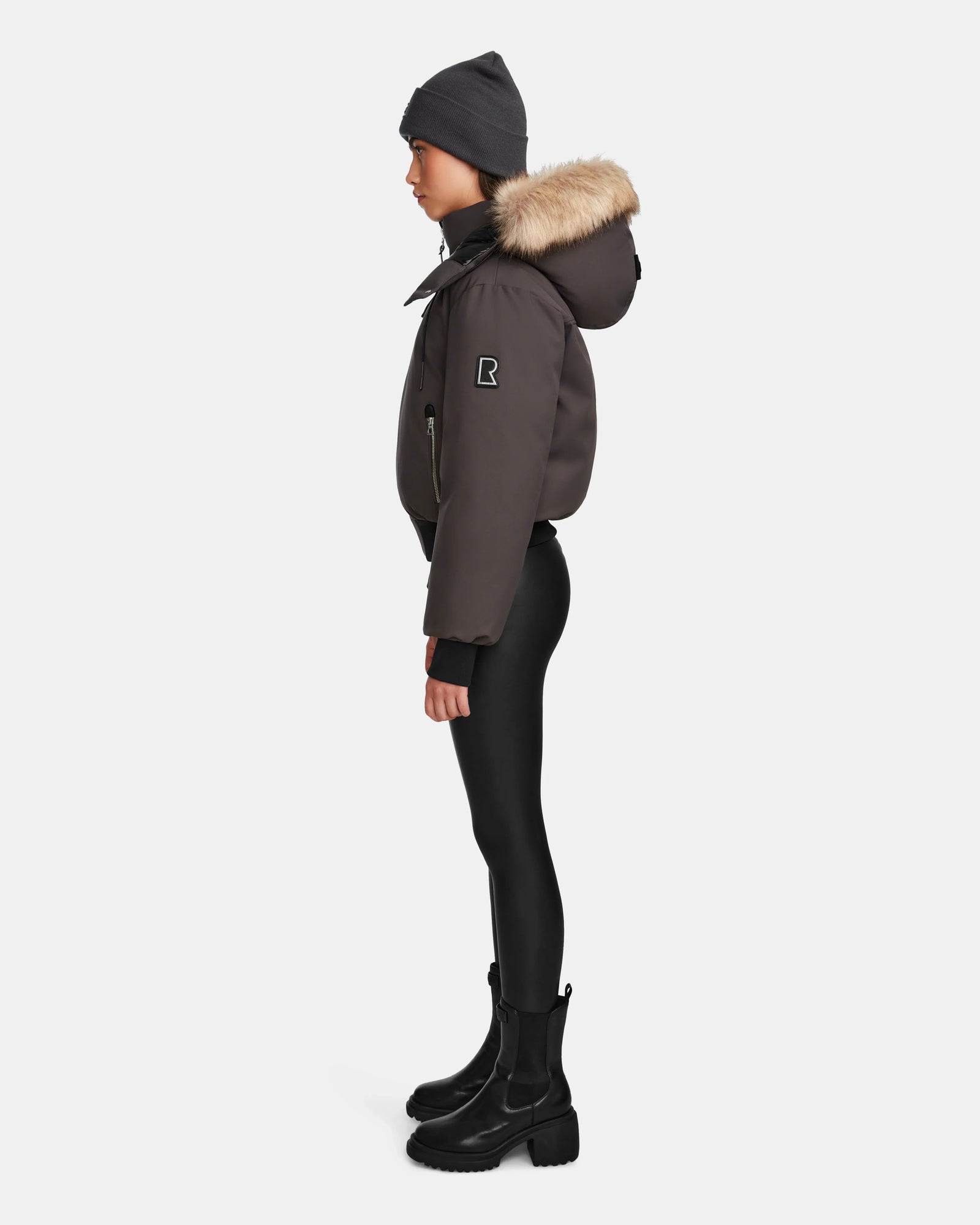 A person stands in profile against a plain white background, wearing the Rudsak MAUDE - L124522 Grey women’s down bomber with fur-lined hood, paired with a gray beanie, black leggings, and black boots.