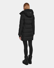 A person with straight, shoulder-length hair is seen from the back wearing the Rudsak MIRIAM - L123544 Black down puffer with a hood, paired with black leggings and boots, standing against a plain light background.