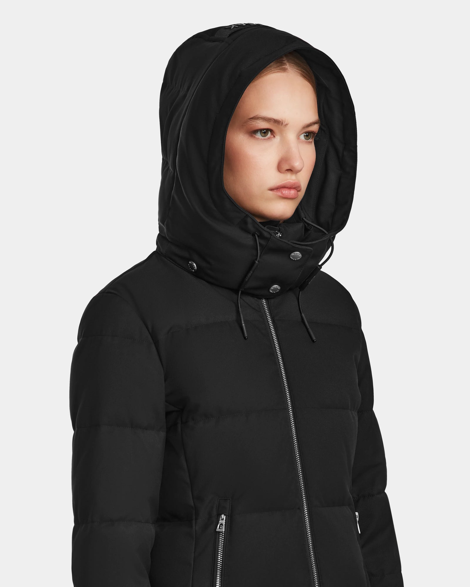 A woman wears the Rudsak MIRIAM - L123544 Black RDS-certified down puffer jacket with hood up, looking forward with a neutral expression against a plain light background.