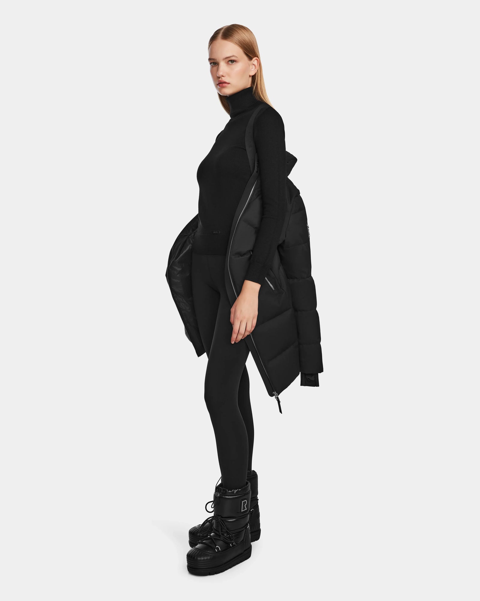 A woman stands sideways against a light background, wearing a black turtleneck, leggings, platform boots, and draping the Rudsak MIRIAM - L123544 Black womens down puffer coat over her shoulder.