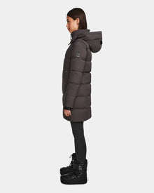 A woman in profile showcases the Rudsak MIRIAM - L123544 Grey down puffer coat with a hood, paired with black leggings and winter boots—highlighting a sleek winter look against a plain white background.