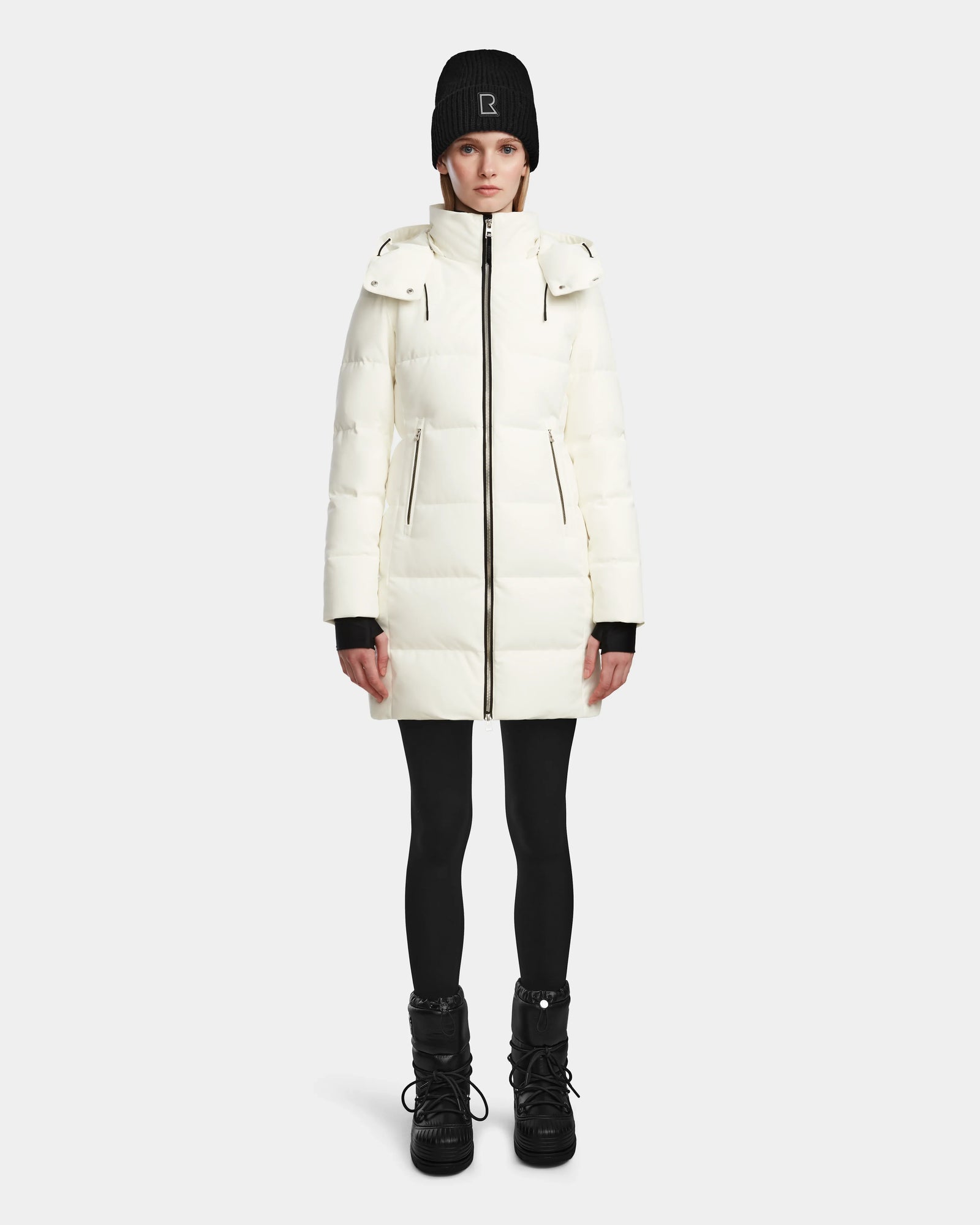 A person wears the Rudsak MIRIAM - L123544 Iceland womens down puffer, paired with a black beanie, black leggings, and black winter boots, standing facing forward against a plain white background.