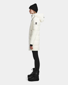 A woman stands in profile against a plain light background wearing the Rudsak MIRIAM - L123544 Iceland womens down puffer with a hood, paired with black leggings, winter boots, and a knit beanie—a stylish and practical cold-weather look.