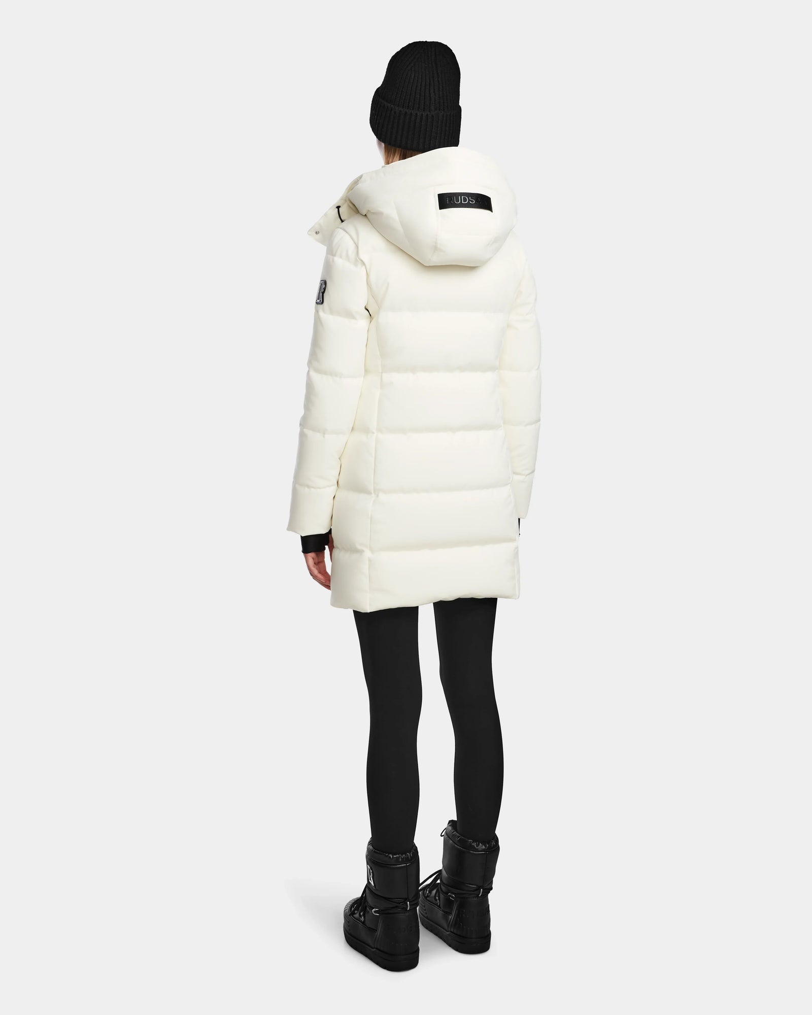 A person wearing the Rudsak MIRIAM - L123544 Iceland womens white down puffer coat with RDS-certified down, black leggings, boots, and a black knit beanie stands facing away from the camera against a plain light background.