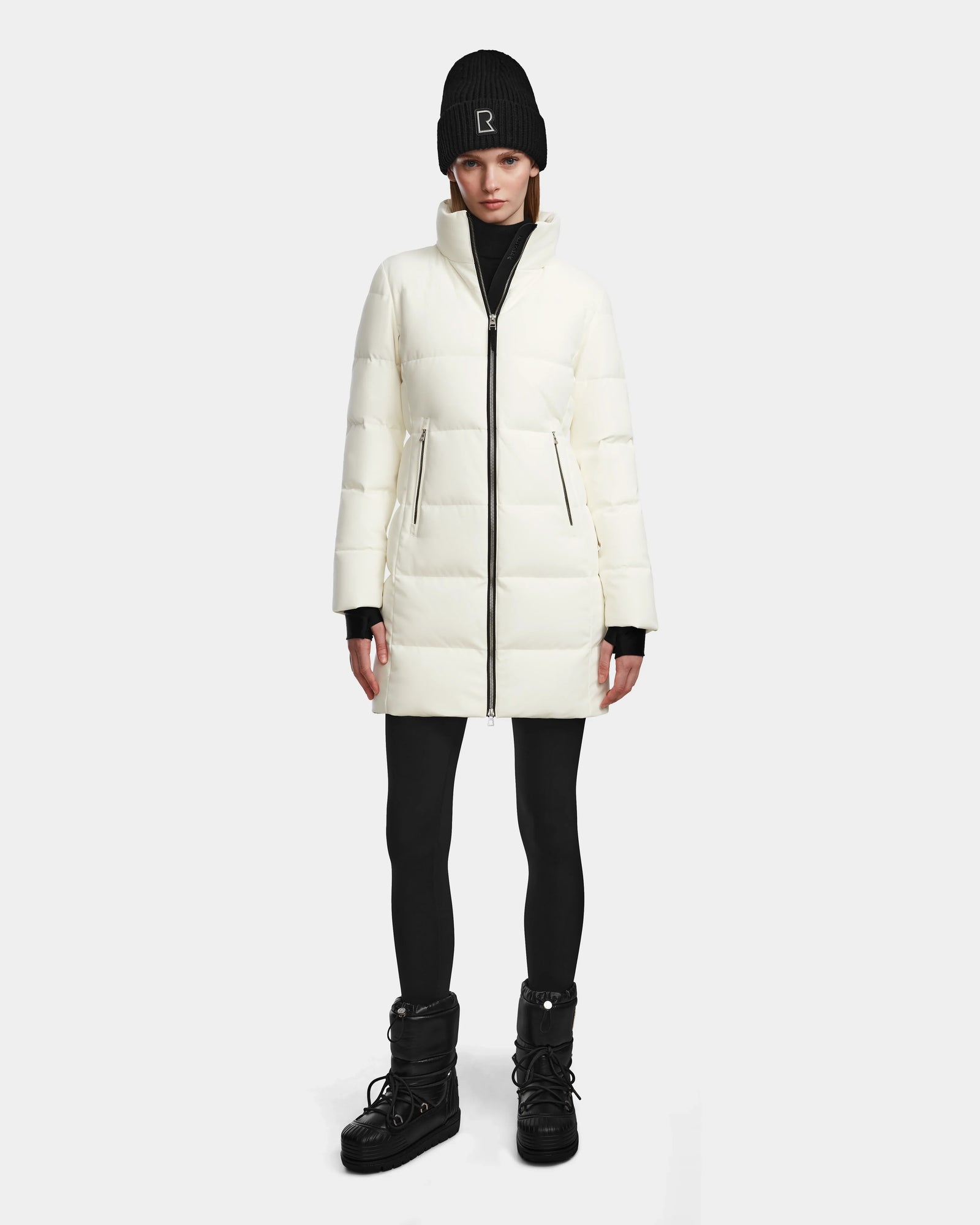 A person wears the Rudsak MIRIAM - L123544 Iceland white women’s down puffer, paired with a black beanie, black leggings, and black winter boots, standing facing forward against a plain light background.